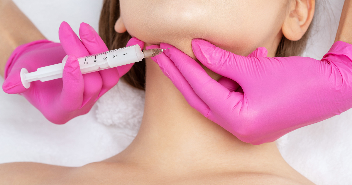 Woman receiving a Kybella injection in her chin area from a medical professional wearing bright pink gloves, using a syringe, in Philadelphia, PA.